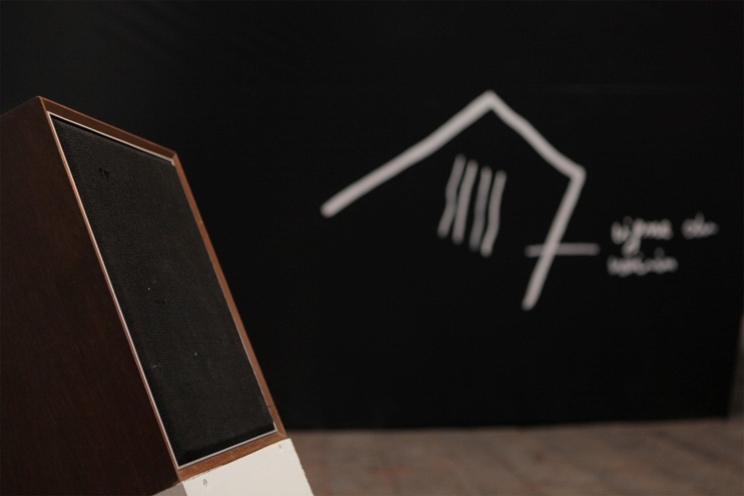 Premier plan : Enceinte sur socle incliné diffusant un recueil de paroles (détail) Arrière plan : Peinture acrylique sur bâche agricole noire (détail)